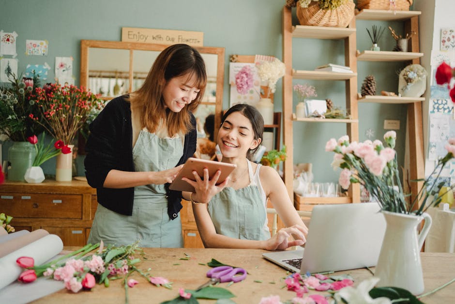 Two women, one standing and the other seated, are inside a flower shop, both smiling and looking at a tablet held by the standing woman. The shop features a wooden counter and shelves filled with vari