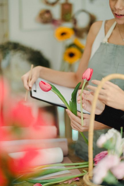 Close-up of a florist's hands holding a vibrant pink tulip with fresh green stems and leaves, while using a tablet to arrange or photograph the flower. The background features a softly blurred floral