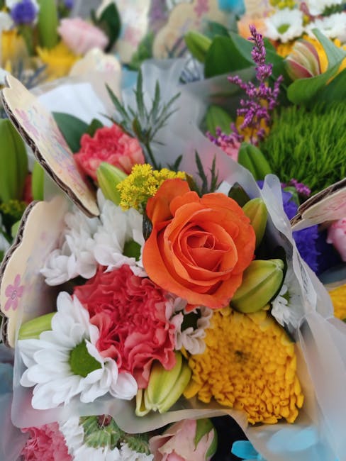 A close-up view of a vibrant floral bouquet arranged with a variety of fresh flowers including a prominent orange rose, pink carnations, white daisies with yellow centers, yellow chrysanthemums, and s