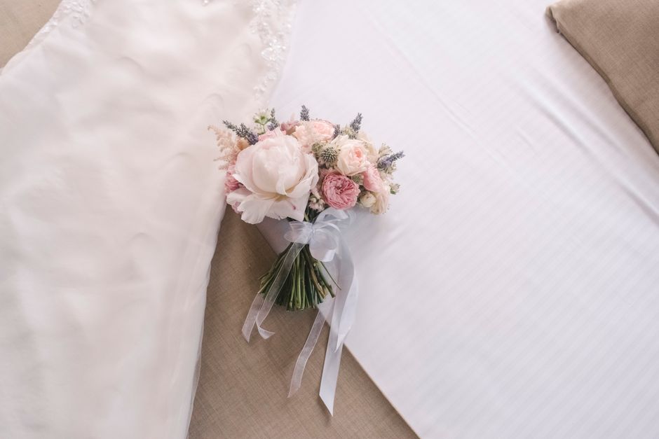 A delicate bouquet of fresh flowers resting on a beige fabric surface, featuring blush pink roses, white peonies, and small lavender sprigs, all wrapped in white tissue paper and tied with a sheer whi
