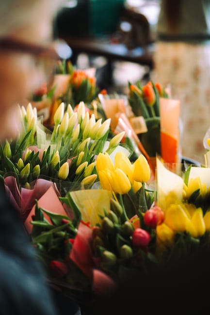 A vibrant display of fresh floral arrangements featuring yellow tulips, white lilies, and pink buds, meticulously wrapped in pastel-colored paper with green foliage accents. The flowers are arranged i