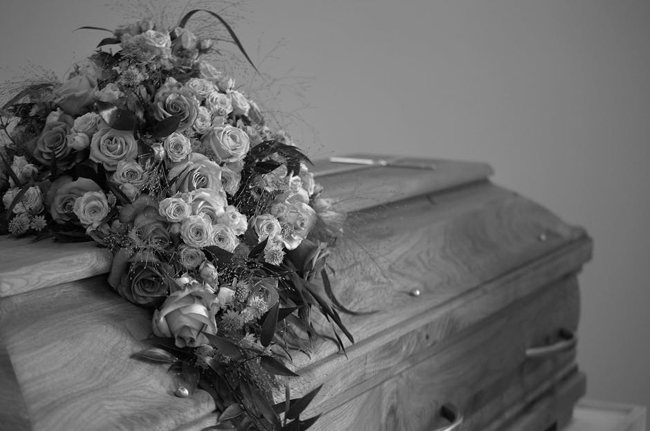 A large bouquet of fresh funeral flowers arranged with cream and deep red roses, white ranunculus, and greenery, resting on the closed lid of a wooden casket. The bouquet is wrapped in dark ribbon and