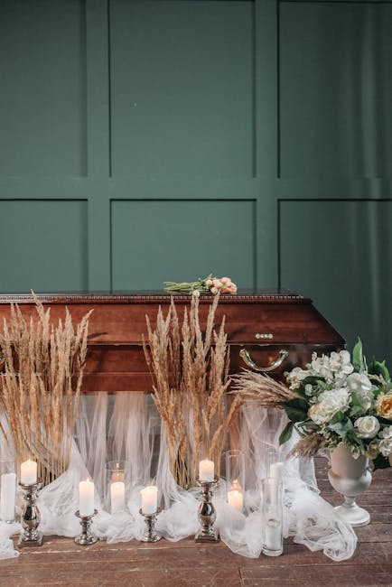 A floral arrangement placed on a wooden piano against a teal paneled wall, featuring white roses, peonies, and greenery in a white ceramic vase. In front of the piano, there are tall dried pampas gras