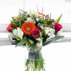Crafted to perfection by an experienced Becontree Heath florist, this breathtaking bouquet is a vibrant testament to the artistry of fresh flower design. Sets of large gerbera daisies-two a radiant and lively shade of orange, two in a soft, inviting peach-stand out with their cheerful presence, while the rich, deep magenta roses nestled nearby add a luxurious undertone. Pristine white alstroemeria and clusters of petite white spray roses meander throughout the ensemble, their multi-petaled forms reminiscent of the gentle blooms found along garden paths near Valence House. Layers of lighter pink blossoms and dainty white fillers create a sense of bountiful abundance, further enhanced by the verdant gloss of deep green foliage. The bouquet's foundation is marked by broad, paddle-shaped aspidistra leaves, gracefully curved to cradle the arrangement, reinforcing its full and rounded silhouette. Slender pointed leaves and delicate greenery fill every niche, drawing the eye and making each flower feel purposefully placed. Set in a sleek, transparent vase, the roots and stems remain visible-symbolic of the bouquet's authenticity and freshness, worthy of any Becontree Heath celebration. Gentle natural light, suggestive of a sun-dappled household or inviting shopfront, caresses the blooms and highlights the colour medley. This display radiates optimism and joy, perfect for birthdays, thank-yous, or simply brightening a home on a Becontree Avenue morning. Every element reflects nature's beauty, curated by local hands and attuned to community spirit.